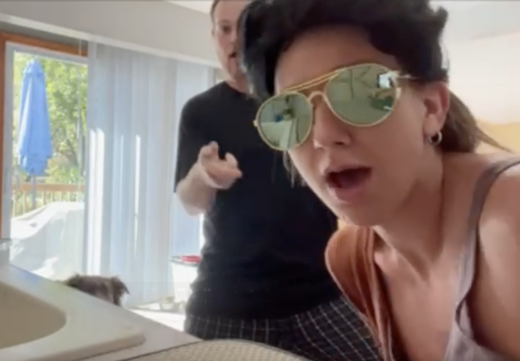 A girl with big gold sunglasses and an elvis wig singing into a camera while someone dances behind her