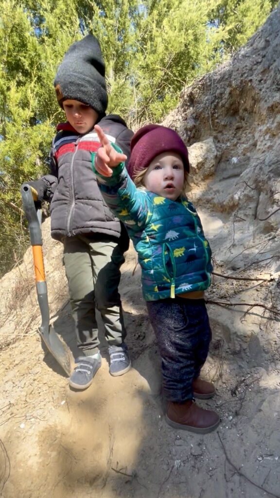 Two young boys outside in winter coats, pants and hats. Standing on the side of a big dirt hill holding a shovel.