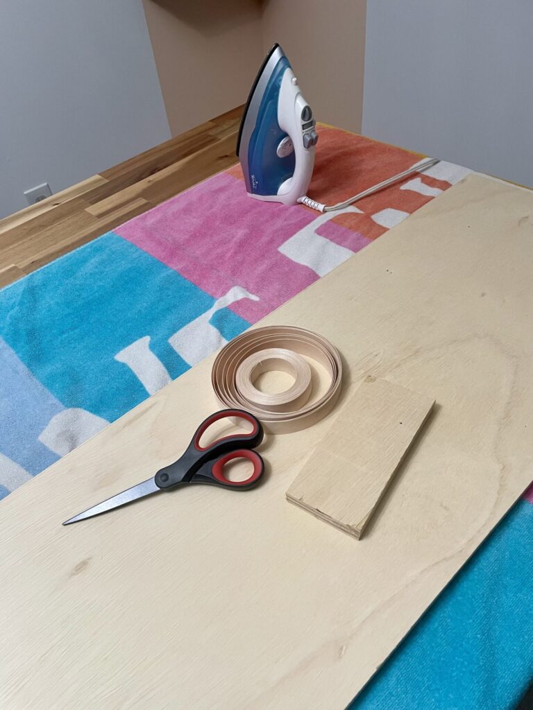 A table with tools for edge banding a piece of plywood. Scissors, and iron, a small piece of scrap wood, and a towel.